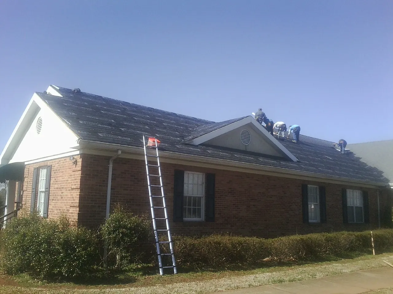 Crew tearing shingles off a brick ranch roof with a ladder set — Metal Roof Repair work by our crew in Blakeslee