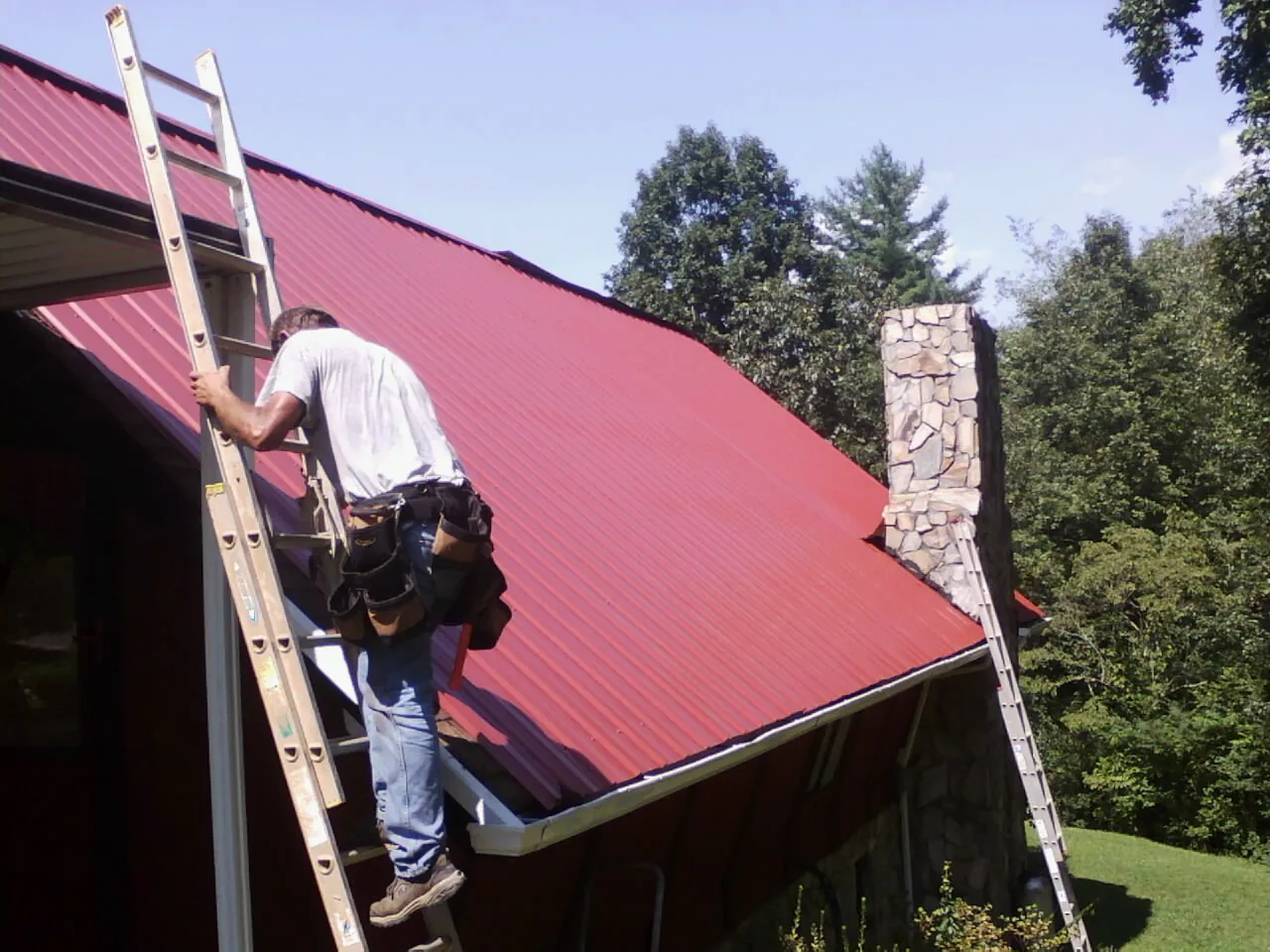 Roofer on a ladder beside a red metal roof and stone chimney — Soffit & Fascia Repair by Roofing Brothers in Blakeslee